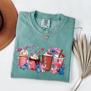 May include: A teal Comfort Colors t-shirt featuring a colorful graphic of cartoon characters and various beverages. The design includes pink, blue, and red elements, with hearts and stars. A gold necklace rests on the shirt.