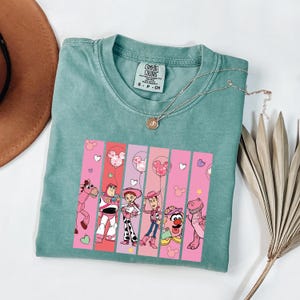 Op de afbeelding: Een teal Comfort Colors t-shirt met een afbeelding van Toy Story-personages. De afbeelding bevat Woody, Buzz Lightyear en Jessie, tegen roze en rode verticale strepen met Mickey Mouse ballonaccenten. Een gouden ketting is over het shirt gedrapeerd.