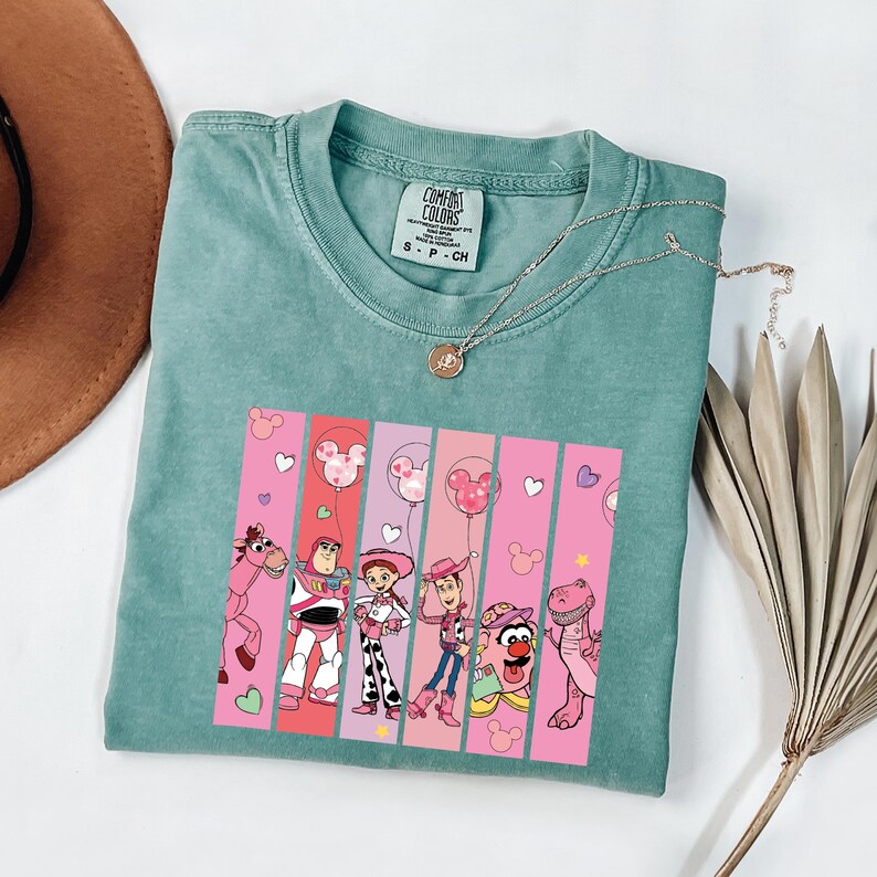 Op de afbeelding: Een teal Comfort Colors t-shirt met een afbeelding van Toy Story-personages. De afbeelding bevat Woody, Buzz Lightyear en Jessie, tegen roze en rode verticale strepen met Mickey Mouse ballonaccenten. Een gouden ketting is over het shirt gedrapeerd.