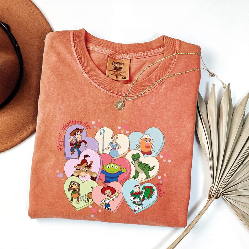 May include: Coral-colored Comfort Colors t-shirt featuring a graphic of Toy Story characters in heart shapes. The phrase "Happy Valentine's Day" is printed on the shirt. A gold necklace and a brown hat are visible in the image.