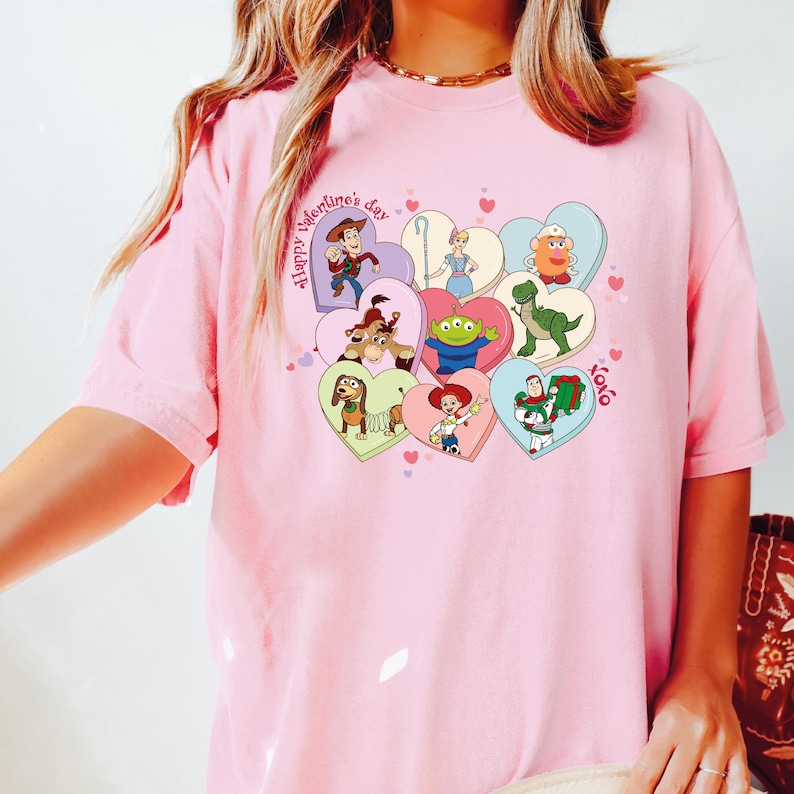 May include: Pink t-shirt featuring a colorful graphic of Toy Story characters inside heart shapes. The phrase "Happy Valentine's Day" is printed on the shirt, along with "xoxo". The design includes Woody, Buzz Lightyear, and other characters.