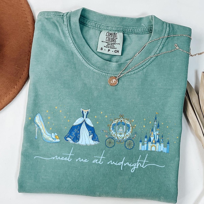 May include: A teal Comfort Colors t-shirt featuring a Cinderella-themed design. The graphic includes a glass slipper, ball gown, carriage, and castle, with the text "meet me at midnight". A silver necklace with a pendant is also visible.