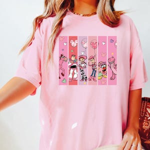 Op de afbeelding: Een lichtroze t-shirt met een kleurrijke afbeelding van stripfiguren in verticale strepen. De afbeelding bevat een roze paard, Buzz Lightyear, Jessie, Woody en een dinosaurus. Mickey Mouse ballonontwerpen zijn ook aanwezig.