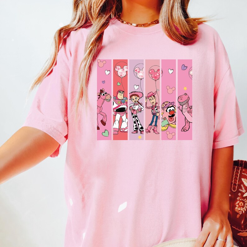 Op de afbeelding: Een lichtroze t-shirt met een kleurrijke afbeelding van stripfiguren in verticale strepen. De afbeelding bevat een roze paard, Buzz Lightyear, Jessie, Woody en een dinosaurus. Mickey Mouse ballonontwerpen zijn ook aanwezig.