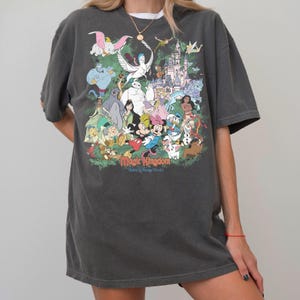 May include: A charcoal gray short-sleeve t-shirt featuring a large, colorful graphic print of various animated characters and a castle. The text "Magic Kingdom" and "Walt Disney World" is printed in a stylized font at the bottom of the graphic.