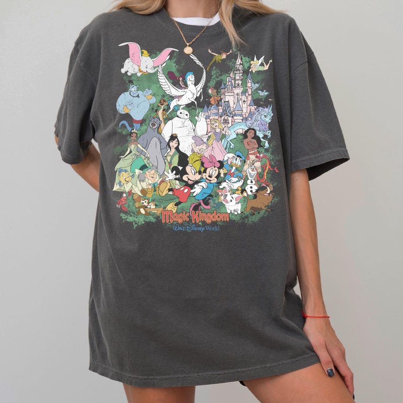 May include: A charcoal gray short-sleeve t-shirt featuring a large, colorful graphic print of various animated characters and a castle. The text "Magic Kingdom" and "Walt Disney World" is printed in a stylized font at the bottom of the graphic.