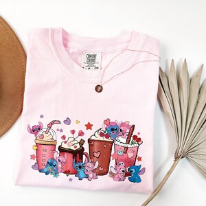 May include: A light pink Comfort Colors t-shirt featuring a graphic of cartoon characters with coffee drinks and hearts. A gold necklace with a pendant is draped on the shirt. A dried palm leaf and a hat are in the background.