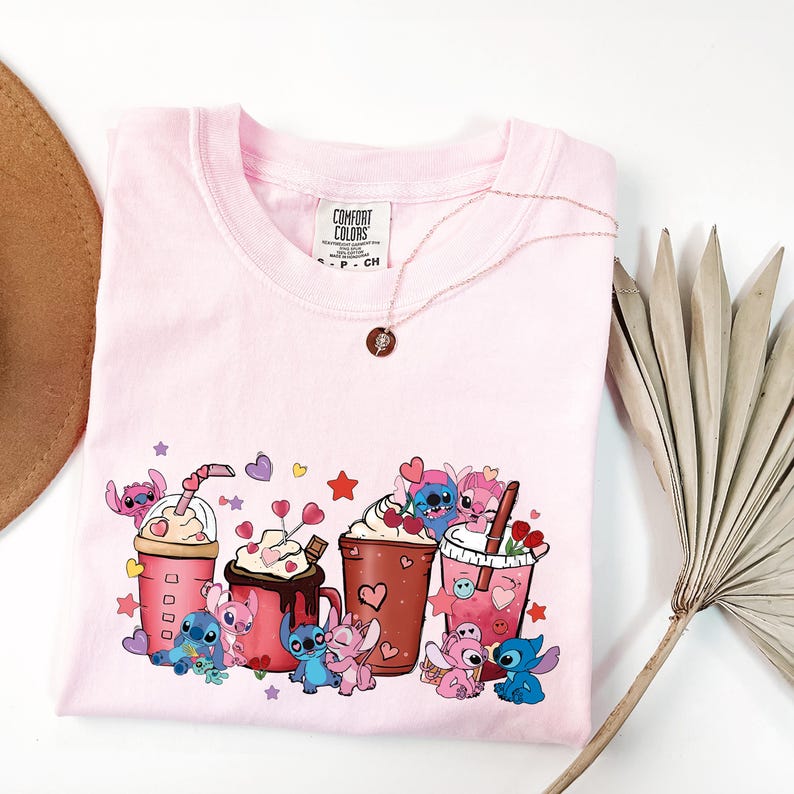 May include: A light pink Comfort Colors t-shirt featuring a graphic of cartoon characters with coffee drinks and hearts. A gold necklace with a pendant is draped on the shirt. A dried palm leaf and a hat are in the background.