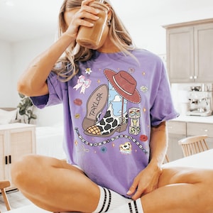 Pu&ograve; includere: Una t-shirt viola a maniche corte con una stampa grafica di uno stivale da cowboy con motivo a macchie di mucca, un cappello marrone, il nome TAYLOR, il numero 13 e blocchi che compongono la scritta BTS. Il design include fiori e un fiocco.