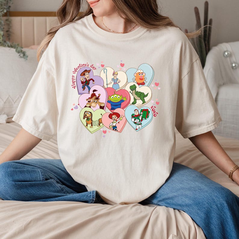 May include: Cream-colored t-shirt featuring a colorful graphic of heart-shaped candies with Toy Story characters. The words "Happy Valentine's Day" and "XOXO" are also printed on the shirt. The shirt is a relaxed fit.