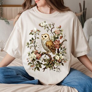 K&ouml;nnte beinhalten: Cremefarbenes T-Shirt mit einer detaillierten Illustration einer Eule auf einem bl&uuml;henden Ast. Die Eule hat braune und wei&szlig;e Federn, umgeben von rosa, wei&szlig;en und gr&uuml;nen floralen Elementen. Das Shirt hat eine lockere Passform.