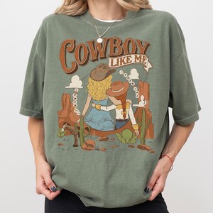 May include: A sage green t-shirt featuring a graphic print of two figures in cowboy hats sitting in a desert landscape with cacti and a horse. The text reads "COWBOY LIKE ME" at the top and "I KNEW YOU" in a circular pattern around the figures.