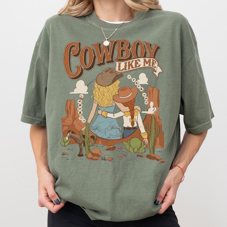May include: A sage green t-shirt featuring a graphic print of two figures in cowboy hats sitting in a desert landscape with cacti and a horse. The text reads "COWBOY LIKE ME" at the top and "I KNEW YOU" in a circular pattern around the figures.