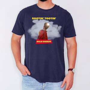 May include: Navy blue t-shirt featuring the text "ROOTIN' TOOTIN'" in yellow above a graphic of a person in a red jacket and the text "BALD SHERIFF" in a red box. The graphic is set against a white cloud.