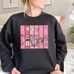 Op de afbeelding: Donkergrijze sweatshirt met een kleurrijke afbeelding van stripfiguren. De afbeelding is verdeeld in verticale panelen met roze en rode achtergronden. De personages zijn onder meer een paard, Buzz Lightyear, Jessie, Woody en anderen. Roze harten en ballonnen zijn ook aanwezig.