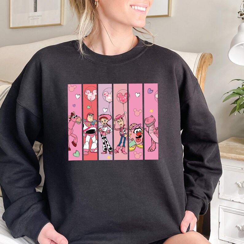 Op de afbeelding: Donkergrijze sweatshirt met een kleurrijke afbeelding van stripfiguren. De afbeelding is verdeeld in verticale panelen met roze en rode achtergronden. De personages zijn onder meer een paard, Buzz Lightyear, Jessie, Woody en anderen. Roze harten en ballonnen zijn ook aanwezig.