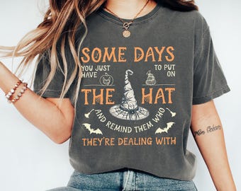 May include: Dark grey t-shirt with orange text that reads "SOME DAYS YOU JUST HAVE TO PUT ON THE HAT AND REMIND THEM WHO THEY'RE DEALING WITH." The graphic includes a witch hat, bats, and a pumpkin.