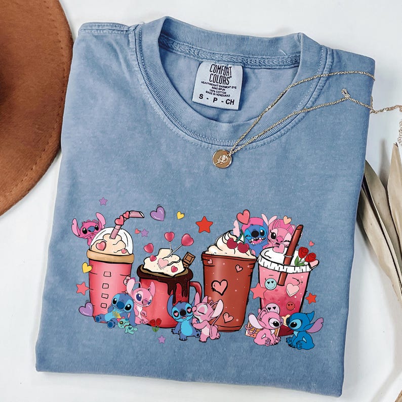May include: A blue Comfort Colors t-shirt featuring a graphic of cartoon characters with various beverages. The design includes pink and blue characters with drinks, hearts, and stars. A gold necklace and a brown hat are also visible.