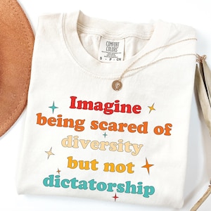 May include: Cream-colored Comfort Colors t-shirt with the text "Imagine being scared of diversity but not dictatorship" in colorful retro font. A gold necklace with a pendant is draped on the shirt. A brown hat is partially visible.