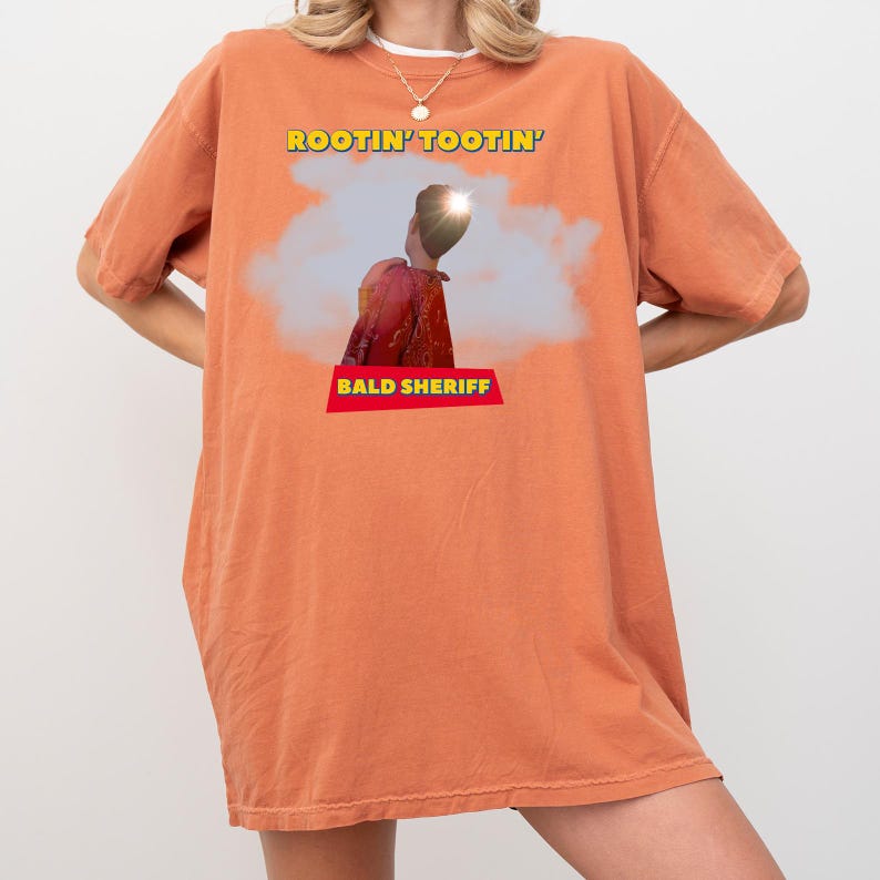 May include: Orange t-shirt featuring the text "ROOTIN' TOOTIN'" above an image of a person in a red shirt and the text "BALD SHERIFF" below. The shirt is a casual style and is orange in colour.