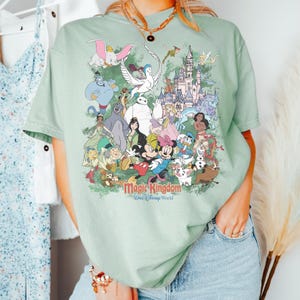 May include: A sage green t-shirt featuring a large, colorful graphic print of various animated characters and a castle. The text "Magic Kingdom" and "Walt Disney World" is printed in red and blue below the characters on the front of the shirt.