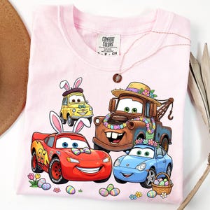 May include: Pink t-shirt featuring cartoon cars decorated with Easter bunny ears and floral garlands. The design includes Lightning McQueen, Mater, and other characters, along with Easter eggs and flowers. The shirt has the Comfort Colors brand tag.
