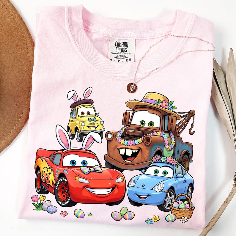 May include: Pink t-shirt featuring cartoon cars decorated with Easter bunny ears and floral garlands. The design includes Lightning McQueen, Mater, and other characters, along with Easter eggs and flowers. The shirt has the Comfort Colors brand tag.