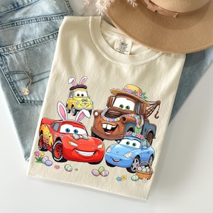 May include: Cream-colored t-shirt featuring cartoon cars decorated for Easter. The graphic includes a red race car, a blue sports car, a brown tow truck, and a yellow car, all with Easter bunny ears and floral decorations. Easter eggs and flowers are scattered around the cars.