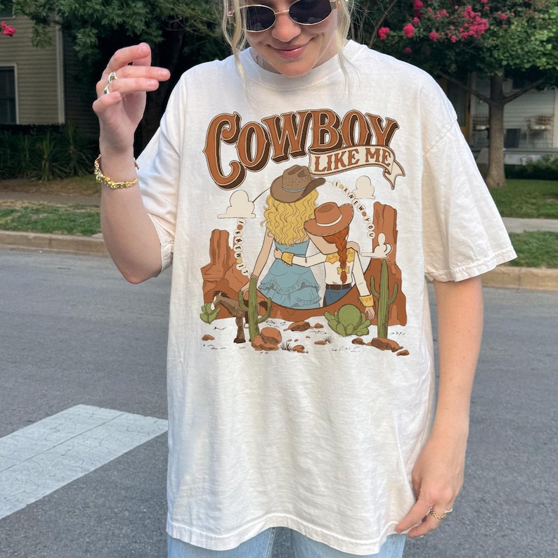 May include: An off-white short-sleeve t-shirt featuring a graphic print with the text "COWBOY LIKE ME" above an illustration of two figures in cowboy hats sitting in a desert landscape with cacti, a horse, and rock formations in shades of brown.