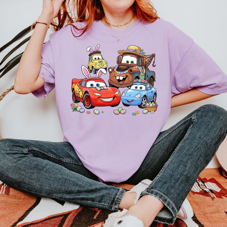 May include: A light purple t-shirt featuring a colorful graphic of animated cars decorated for Easter. The cars include a red race car, a blue car, a brown tow truck, and a yellow car, all with Easter bunny ears and surrounded by Easter eggs.