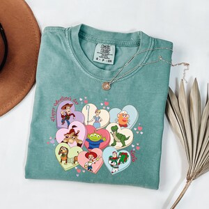 May include: A teal Comfort Colors t-shirt featuring a heart-shaped graphic with various Toy Story characters. The graphic includes the text "Happy Valentine's Day" and "XOXO". A delicate silver necklace rests on the shirt.