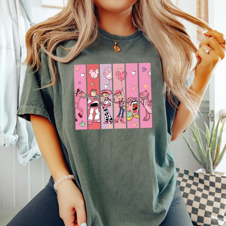 Op de afbeelding: Donkergroen T-shirt met een kleurrijke afbeelding van stripfiguren in verticale strepen. De afbeelding bevat personages als Woody en Buzz Lightyear, met roze en witte accenten. Het shirt is een casual, comfortabele stijl.