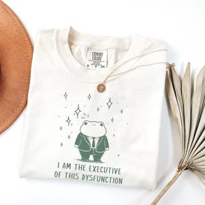 May include: Cream-colored t-shirt featuring a cartoon frog in a suit, with the text "I AM THE EXECUTIVE OF THIS DYSFUNCTION." A gold necklace and a brown hat are also visible. The shirt has a Comfort Colors tag.