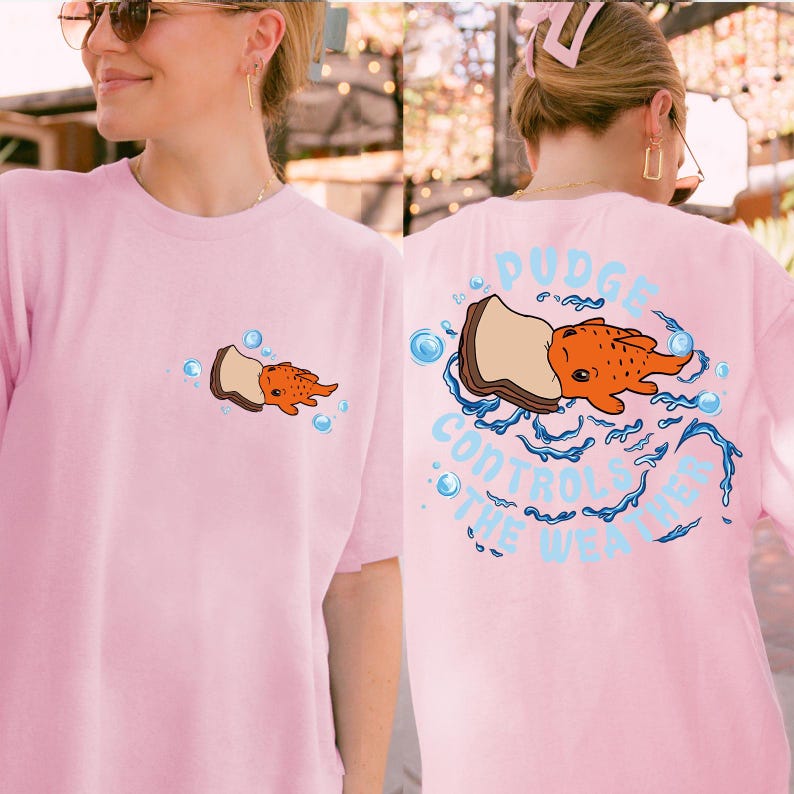 K&ouml;nnte beinhalten: Hellrosa T-Shirt mit einem Cartoon-Design eines orangefarbenen Fisches und einem Brotst&uuml;ck. Der Text auf der R&uuml;ckseite des Shirts lautet "Pudge Controls The Weather" in Hellblau. Blasen und Wassergrafiken umgeben den Fisch.