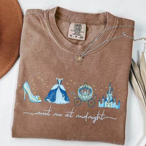 May include: Brown Comfort Colors t-shirt with a graphic of a blue high heel, ball gown, carriage, and castle. The text "meet me at midnight" is written in cursive below the graphic. A silver necklace with a small pendant is on the shirt.