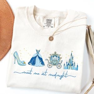 May include: Cream-colored t-shirt featuring a blue illustration of a glass slipper, ball gown, carriage, and castle. The text "meet me at midnight" is written in blue script. A gold necklace with a pendant is draped across the shirt.