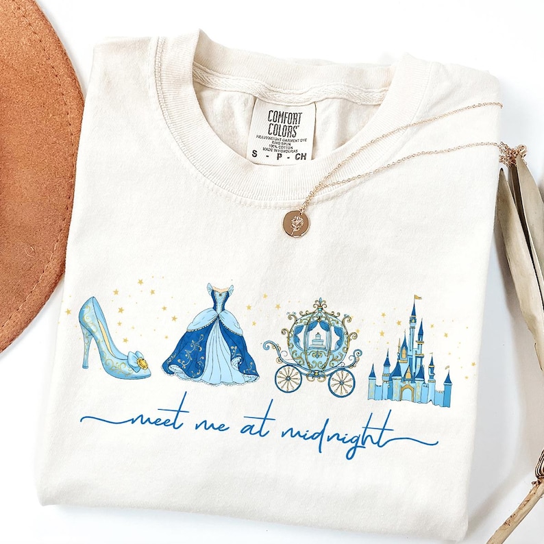 May include: Cream-colored t-shirt featuring a blue illustration of a glass slipper, ball gown, carriage, and castle. The text "meet me at midnight" is written in blue script. A gold necklace with a pendant is draped across the shirt.
