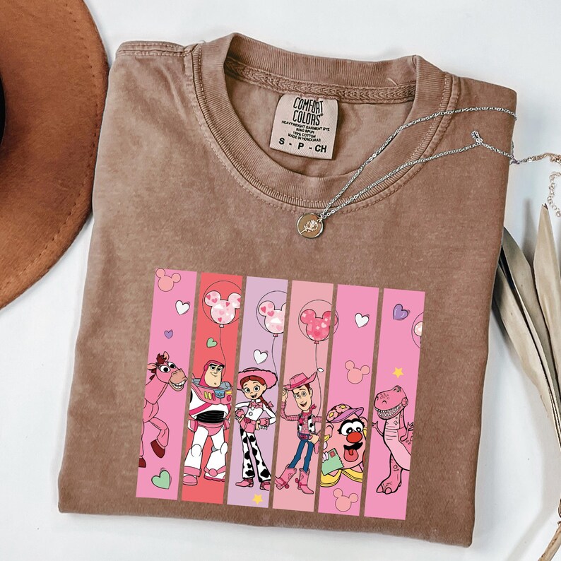 Op de afbeelding: Bruin T-shirt met een afbeelding van Toy Story-personages. De afbeelding bevat Buzz Lightyear, Woody, Jessie en anderen, tegen een roze en witte gestreepte achtergrond met Mickey Mouse ballonaccenten. Een zilveren ketting ligt over het shirt.