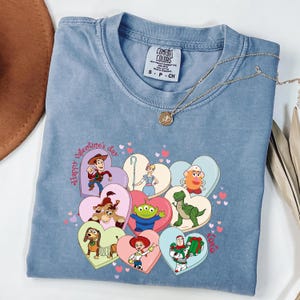 May include: A blue Comfort Colors t-shirt featuring a graphic of Toy Story characters in heart-shaped designs. The shirt has the text "Happy Valentine's Day" and a gold necklace.