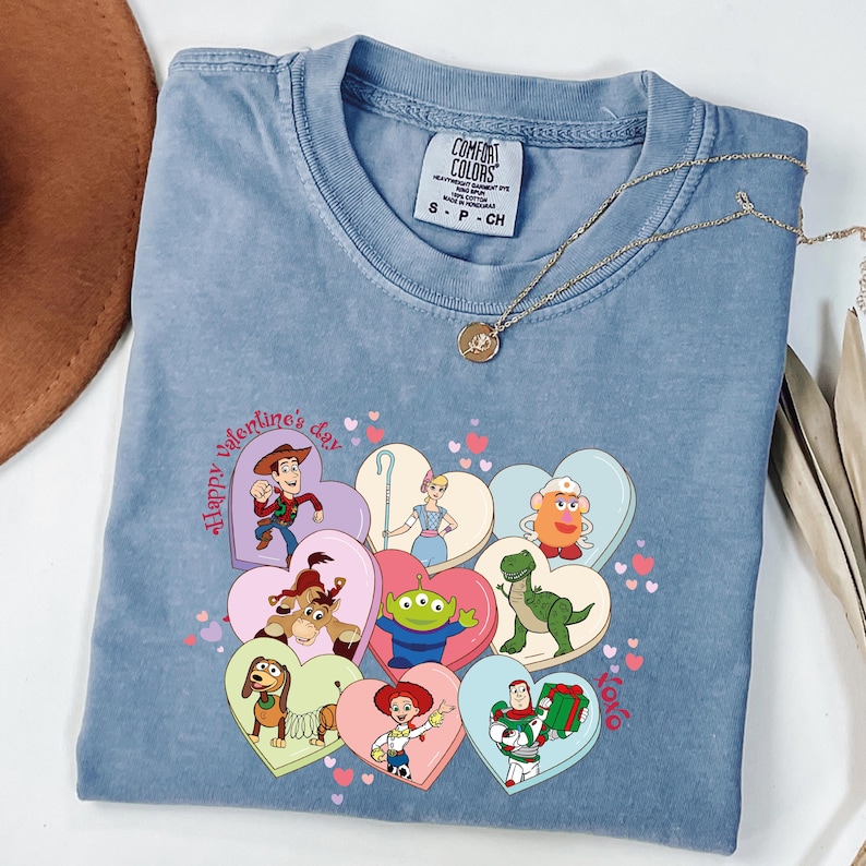 May include: A blue Comfort Colors t-shirt featuring a graphic of Toy Story characters in heart-shaped designs. The shirt has the text "Happy Valentine's Day" and a gold necklace.