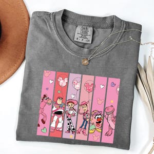 Op de afbeelding: Grijze Comfort Colors t-shirt met een afbeelding van Toy Story-personages op roze en rode verticale strepen. Het ontwerp bevat Woody, Buzz Lightyear, Jessie en andere personages met Mickey Mouse ballonaccenten.