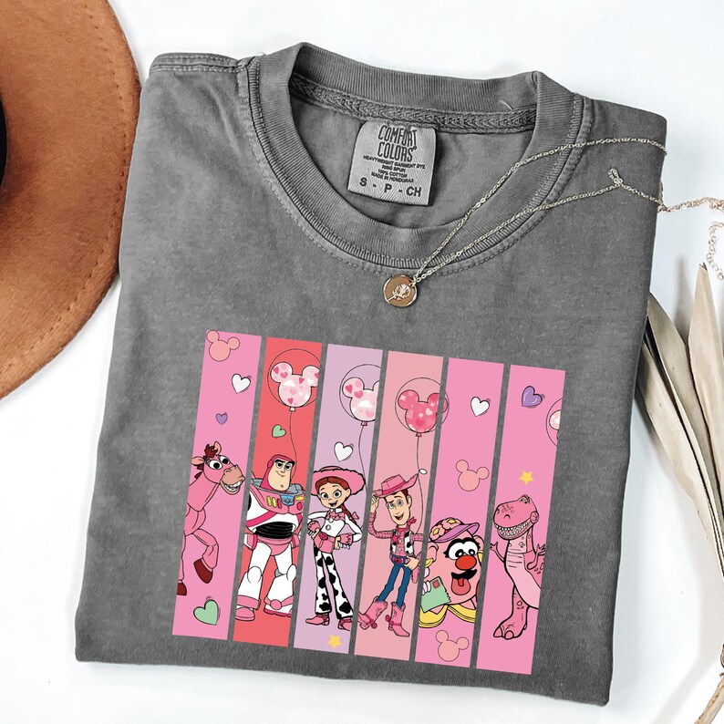 Op de afbeelding: Grijze Comfort Colors t-shirt met een afbeelding van Toy Story-personages op roze en rode verticale strepen. Het ontwerp bevat Woody, Buzz Lightyear, Jessie en andere personages met Mickey Mouse ballonaccenten.