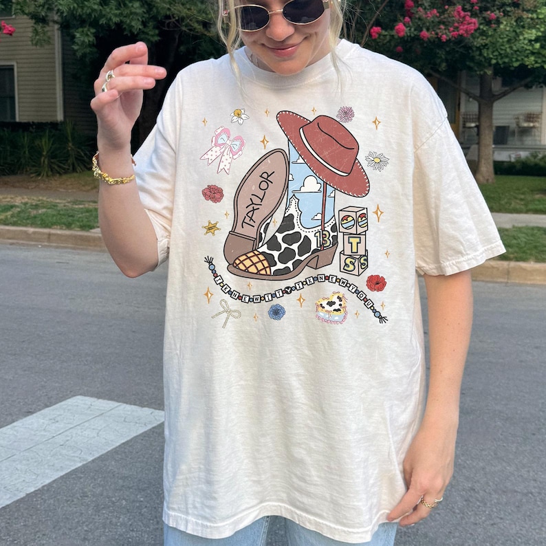 Pu&ograve; includere: Una t-shirt a maniche corte color bianco sporco con una stampa grafica raffigurante uno stivale da cowboy maculato con il nome TAYLOR, un cappello da cowboy marrone, blocchi con la scritta BTS, una catenella di perline e fiori e stelle.