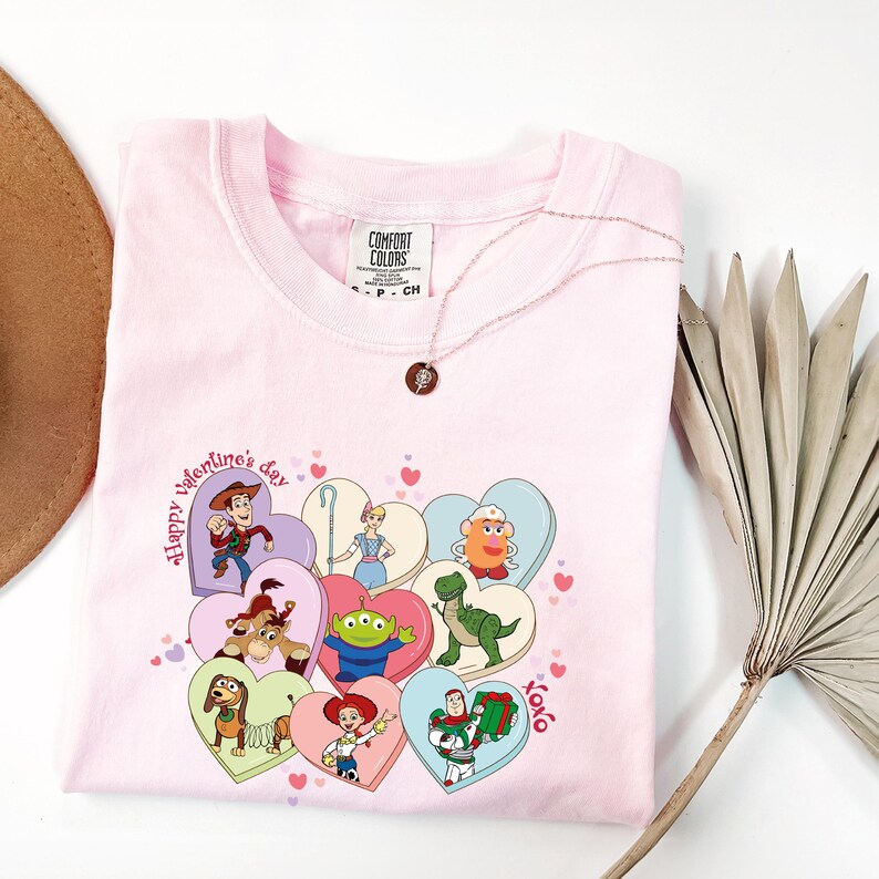 May include: A light pink t-shirt featuring a graphic of Toy Story characters on heart-shaped designs. The words "Happy Valentine's Day" and "Xoxo" are also printed on the shirt. A gold necklace with a pendant is draped on the shirt.