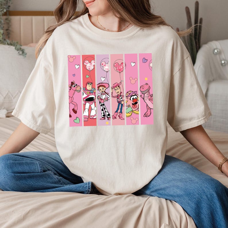 Op de afbeelding: Cr&egrave;mekleurig T-shirt met een kleurrijke afbeelding van stripfiguren in roze en rode verticale strepen. De afbeelding bevat een roze paard, Buzz Lightyear, Jessie, Woody, een Muppet en een dinosaurus. Er zijn ook harten en Mickey Mouse silhouetten.