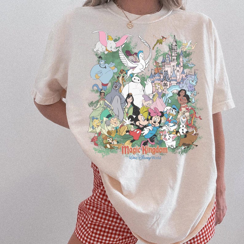May include: A cream-colored short-sleeve t-shirt featuring a large, colorful graphic print of various animated characters and a castle. The text "Magic Kingdom" and "Walt Disney World" is printed in red and blue below the character illustration.