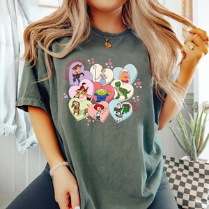 May include: Dark green t-shirt featuring a colorful graphic of cartoon characters in heart shapes. The phrase "Happy Valentine's Day" is printed above the hearts. The shirt is made of a soft material.