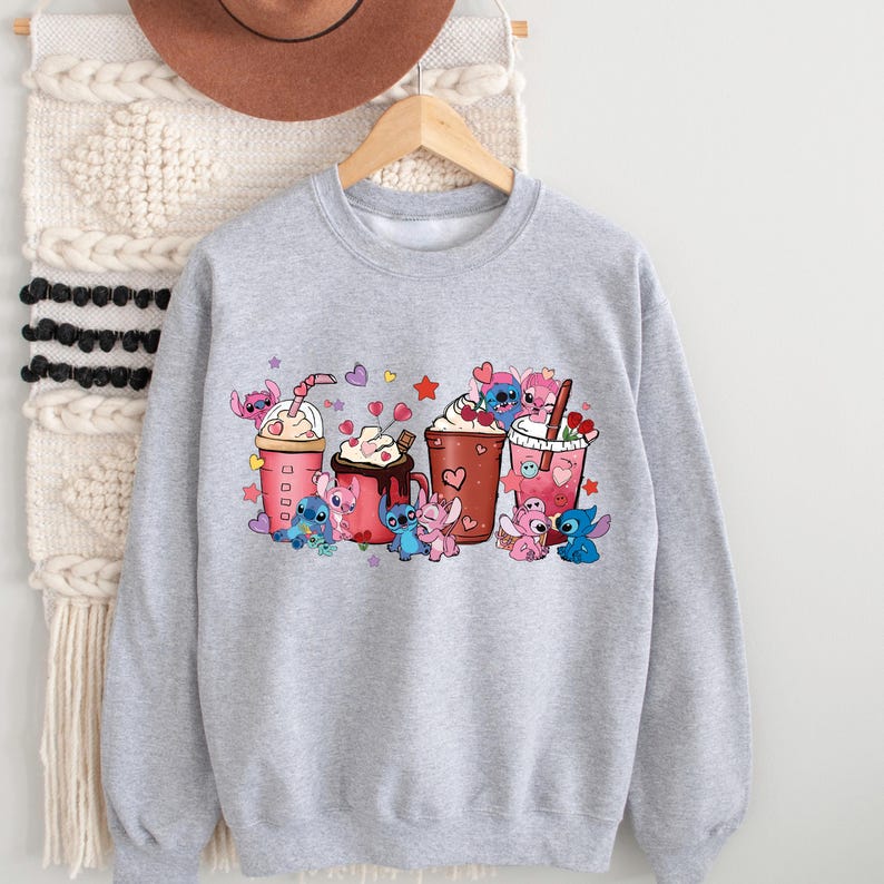 May include: A light gray sweatshirt featuring a colorful graphic of cartoon characters with various beverages. The graphic includes pink, red, and blue characters with hearts and stars. The sweatshirt has a crew neck and long sleeves.