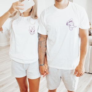 Puede incluir: Dos personas con camisetas blancas a juego con dise&ntilde;os de personajes de dibujos animados. Una camiseta presenta a Daisy Duck, la otra a Donald Duck, ambas en contorno morado. Tambi&eacute;n llevan pantalones cortos grises.