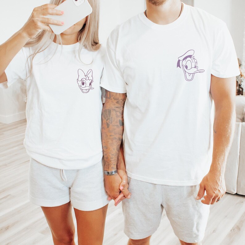 Puede incluir: Dos personas con camisetas blancas a juego con dise&ntilde;os de personajes de dibujos animados. Una camiseta presenta a Daisy Duck, la otra a Donald Duck, ambas en contorno morado. Tambi&eacute;n llevan pantalones cortos grises.